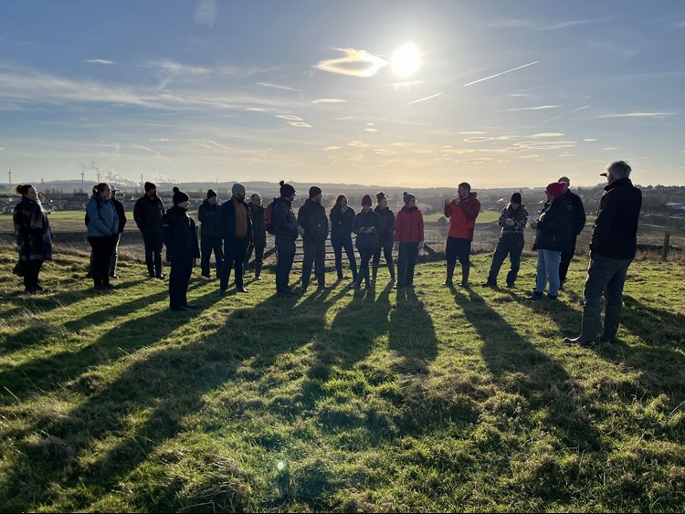 A group of around 20 people listening to a man talk, standing in a field.